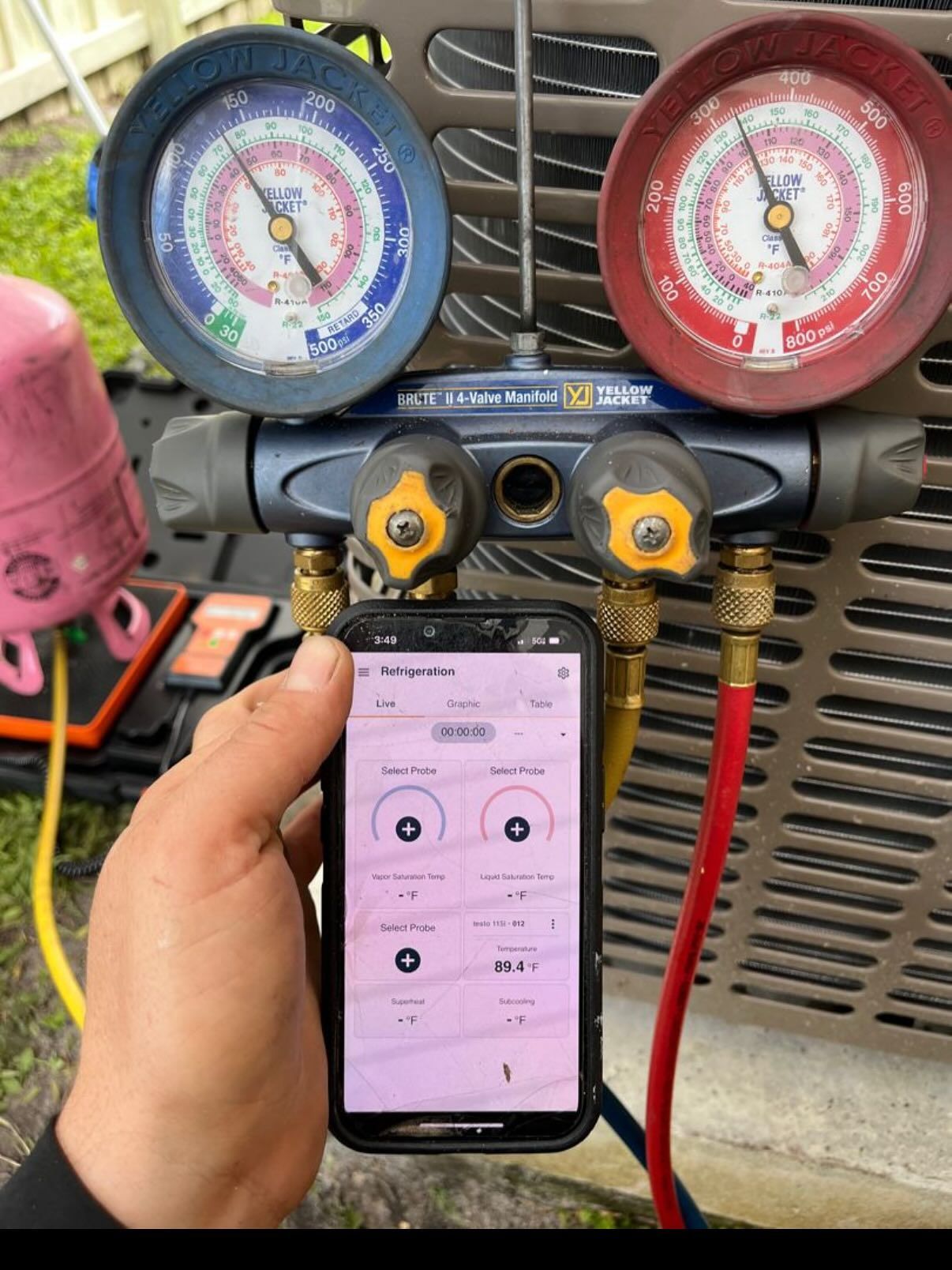 A technician’s hand holding a phone displaying HVAC diagnostic data alongside analog gauges connected to an AC unit.