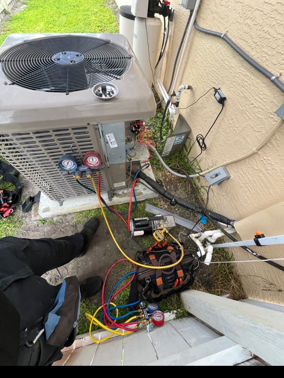 A technician's hands and legs visible while servicing an outdoor air conditioning unit with a manifold gauge and vacuum pump.