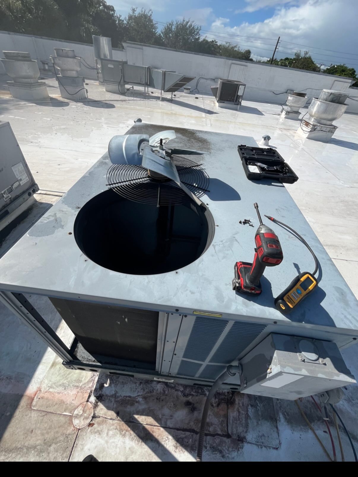 A rooftop HVAC unit with a removed fan motor, a power drill, and testing equipment placed on its surface.