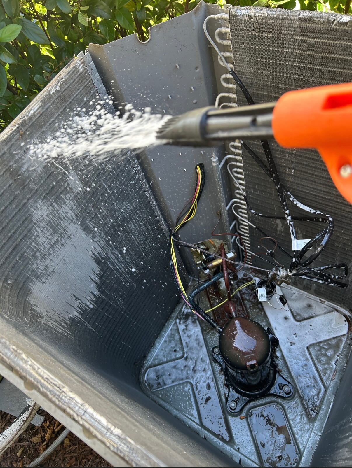 A person uses a hose nozzle to spray water onto the metal cooling coils of an outdoor air conditioning unit.