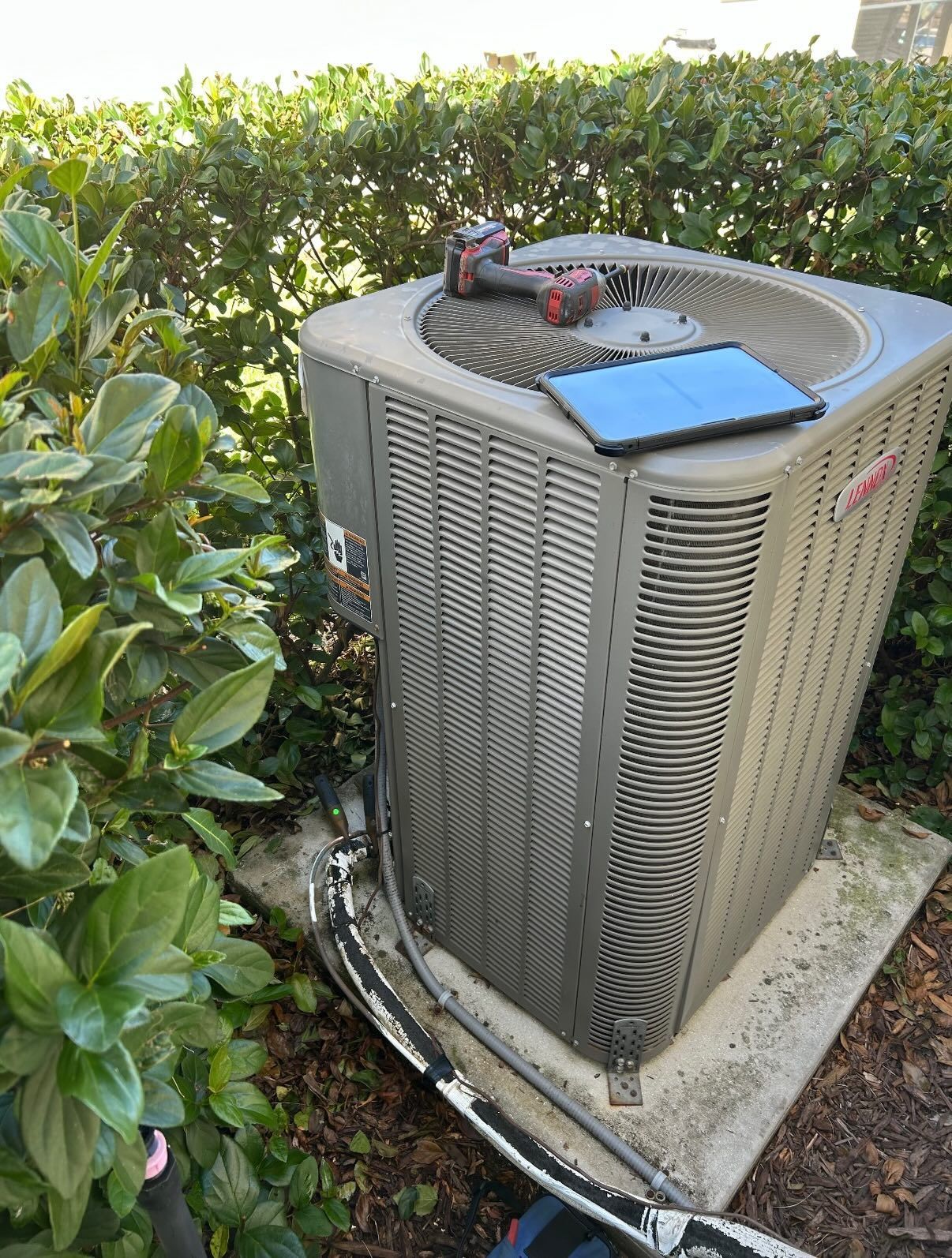 An outdoor HVAC unit sitting on a concrete pad next to a hedge, with a power tool and a tablet placed on its top grille.