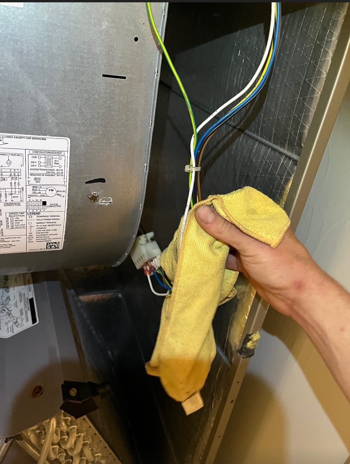 A hand holds a yellow microfiber cloth while cleaning the interior wiring and metal housing of an HVAC unit.