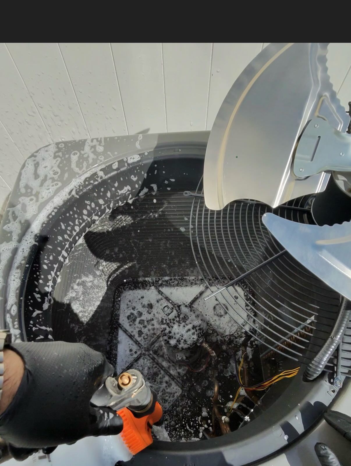 A gloved hand spraying cleaning foam into the open, dark interior of an outdoor air conditioning unit fan compartment.
