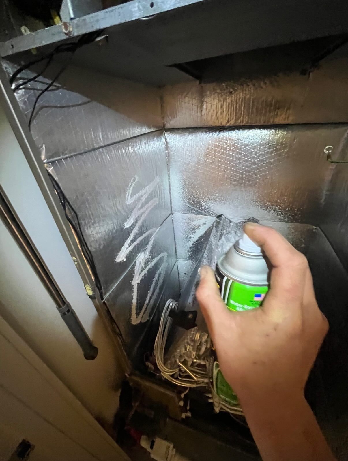 A person sprays cleaner inside an HVAC unit, targeting the metal coils inside the silver-insulated cabinet.