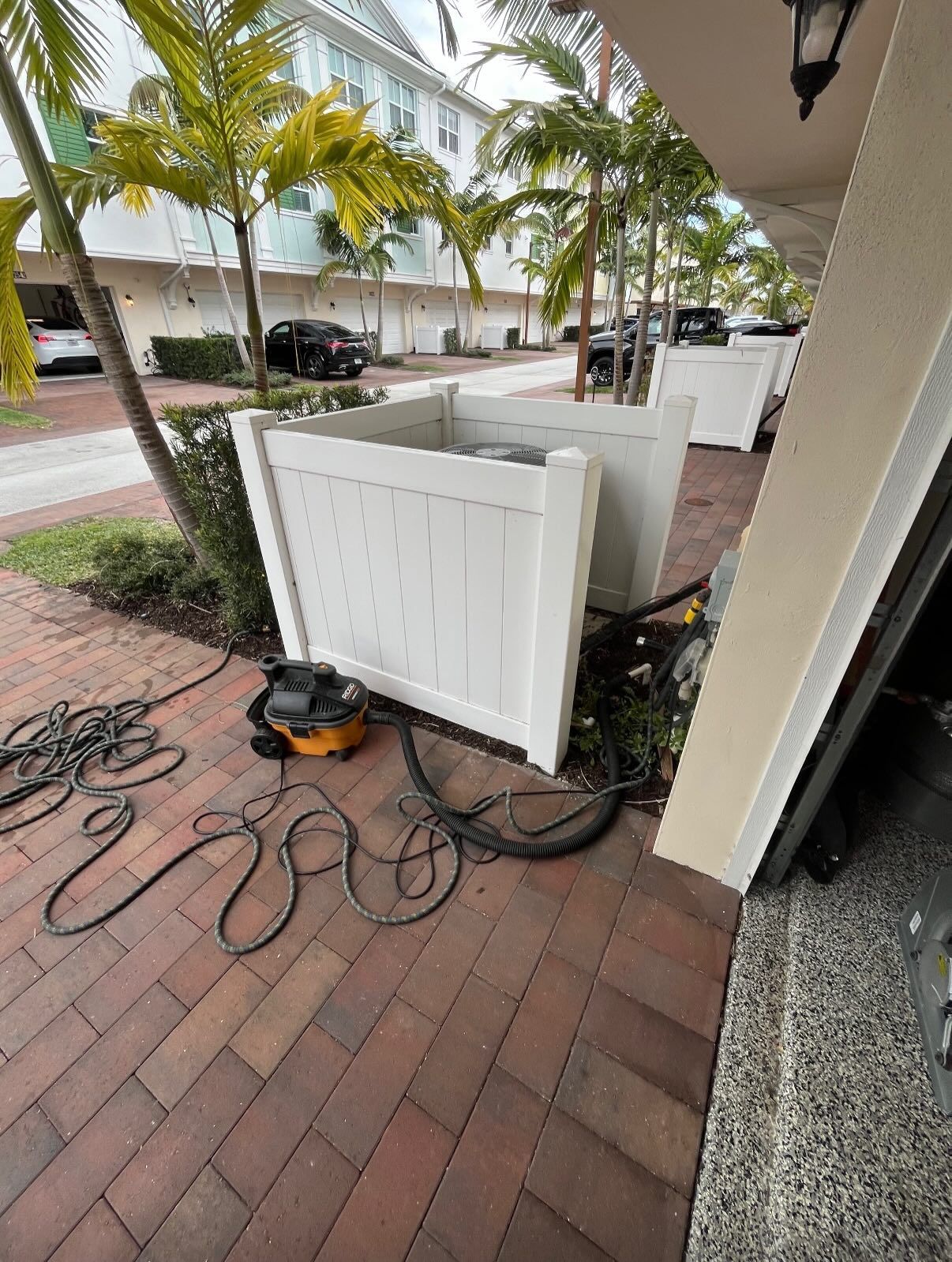 A white fence encloses an AC unit on a brick patio with a shop vacuum and power cables lying on the ground nearby.