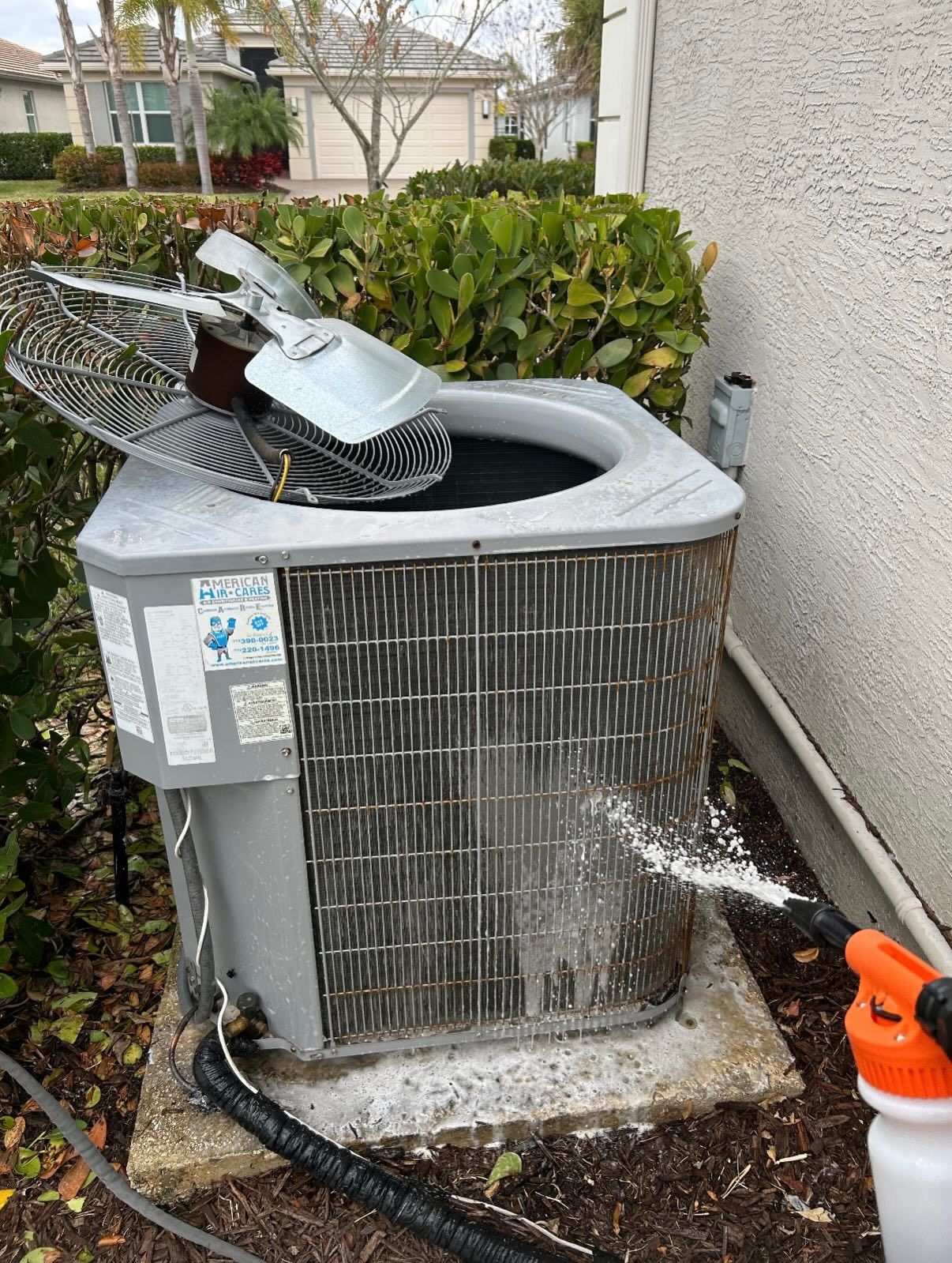 A person uses a spray bottle to clean the metal condenser coils of an outdoor residential HVAC unit.