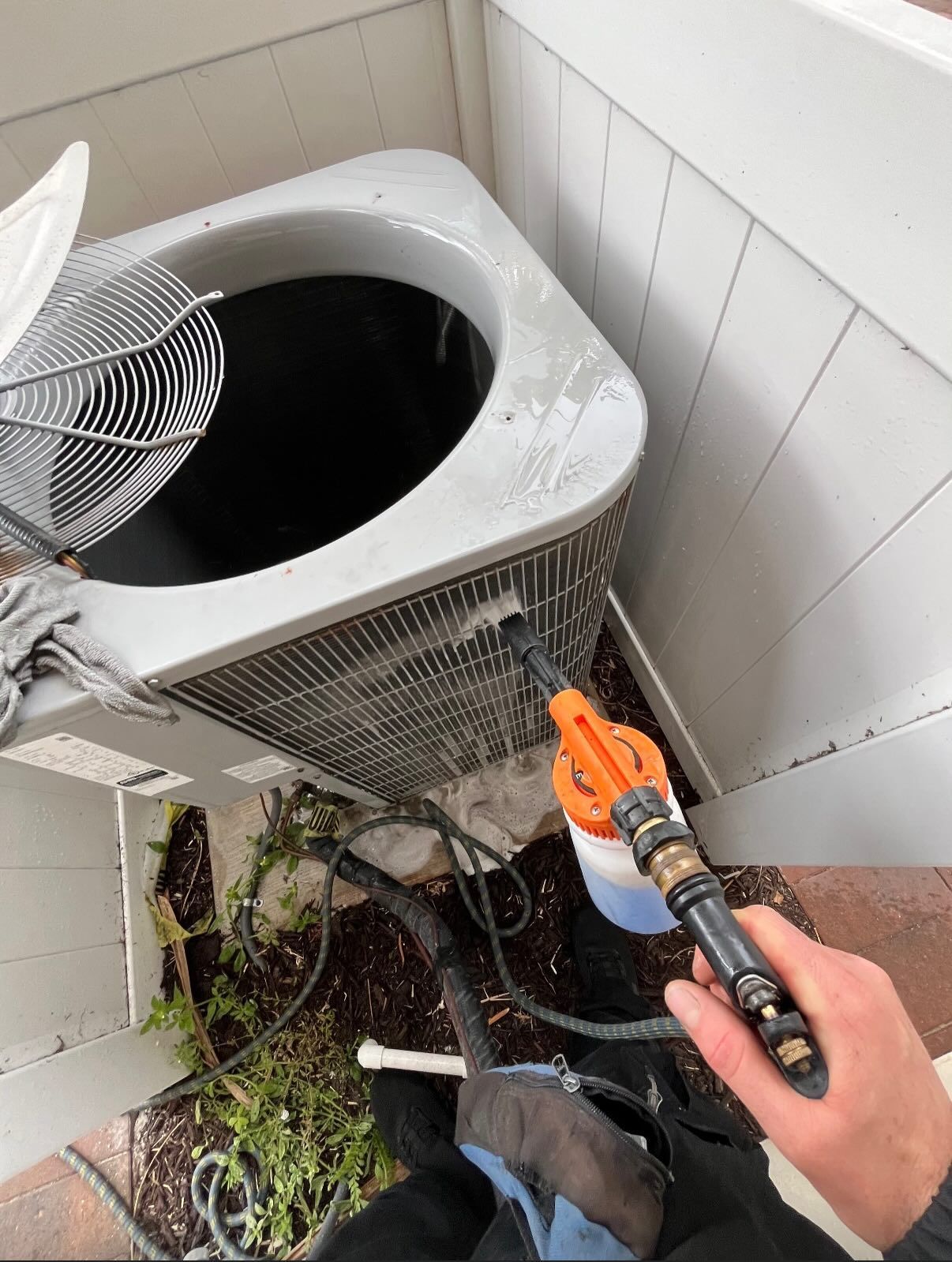 A person sprays cleaning foam onto the exterior condenser coils of an outdoor air conditioning unit.