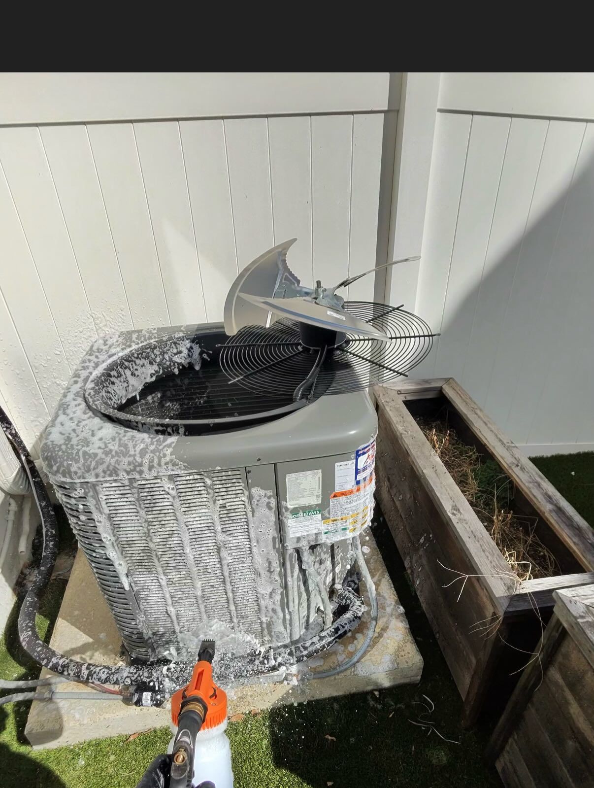 A person uses a spray bottle to apply white cleaning foam to the exterior coils of an outdoor air conditioning unit.