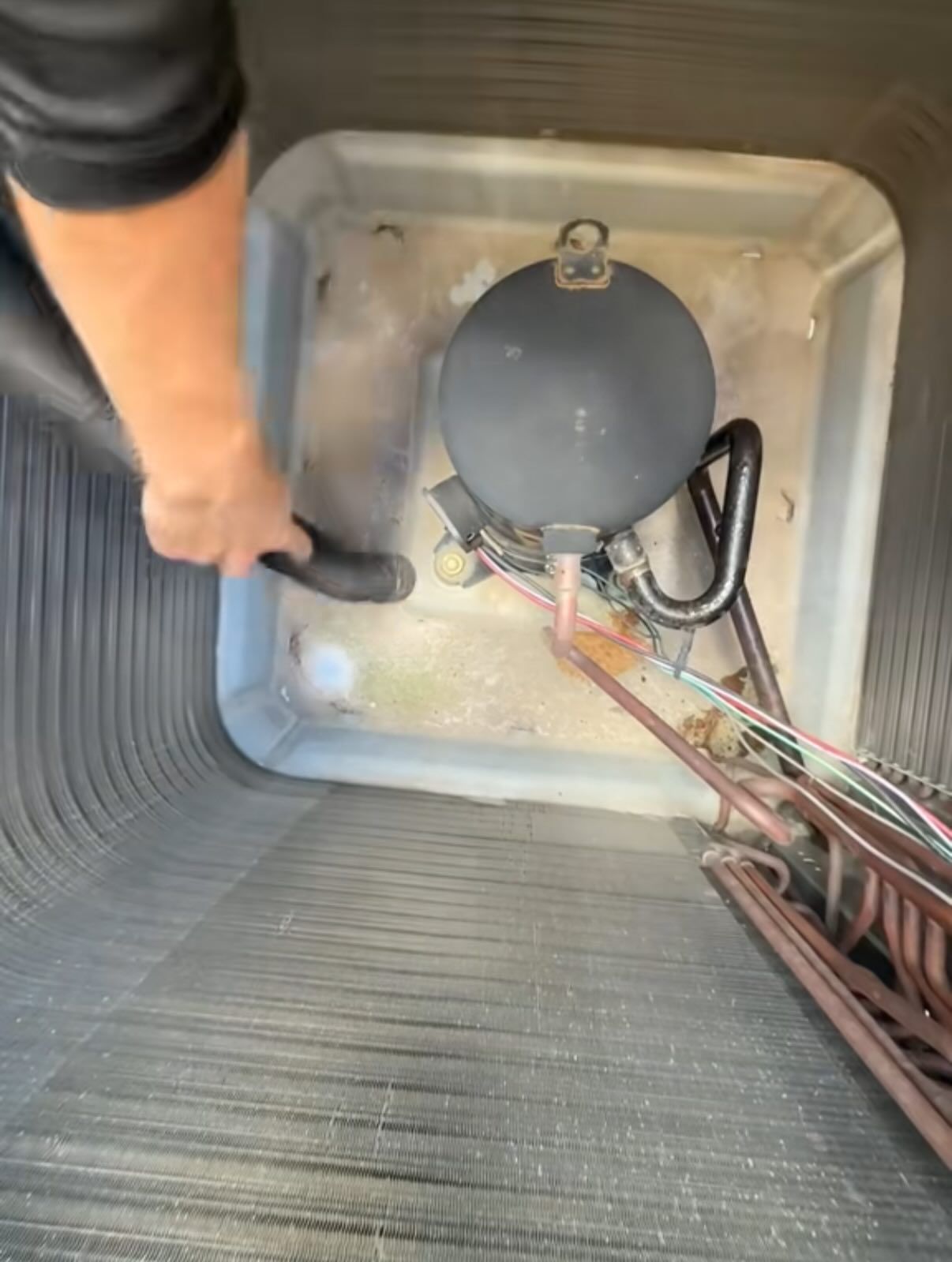 A person using a vacuum hose to clean debris from the bottom of an open outdoor air conditioning unit.