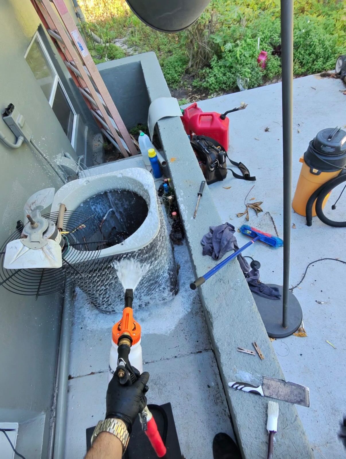 A person wearing gloves sprays foaming cleaner onto the outdoor fan coil of an air conditioning unit.