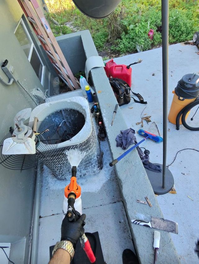 A person wearing gloves sprays foaming cleaner onto the outdoor fan coil of an air conditioning unit.