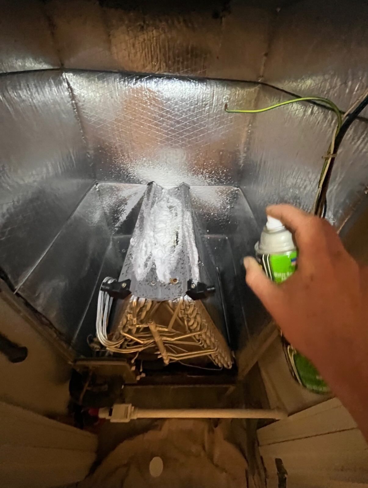 A hand sprays cleaner into an open HVAC duct housing containing metal coils and silver insulation lining.