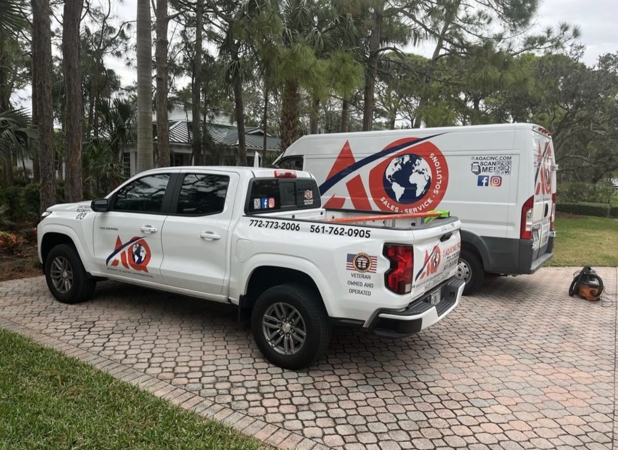 A white pickup truck and a white work van with AC branding parked on a paved driveway in front of a tree-lined house.