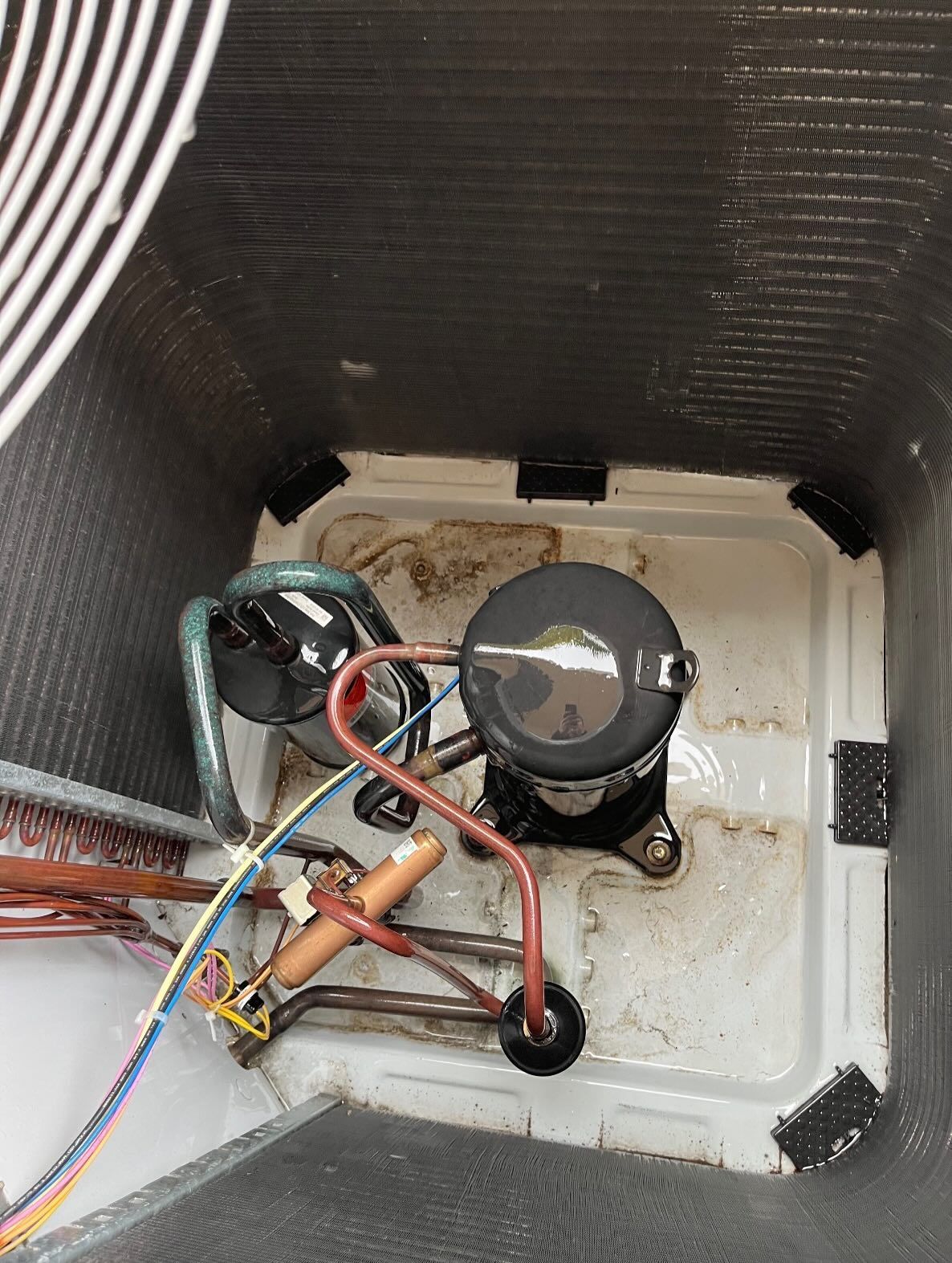 An overhead view of an open outdoor HVAC unit, showing a black compressor, copper piping, and debris in the white base.