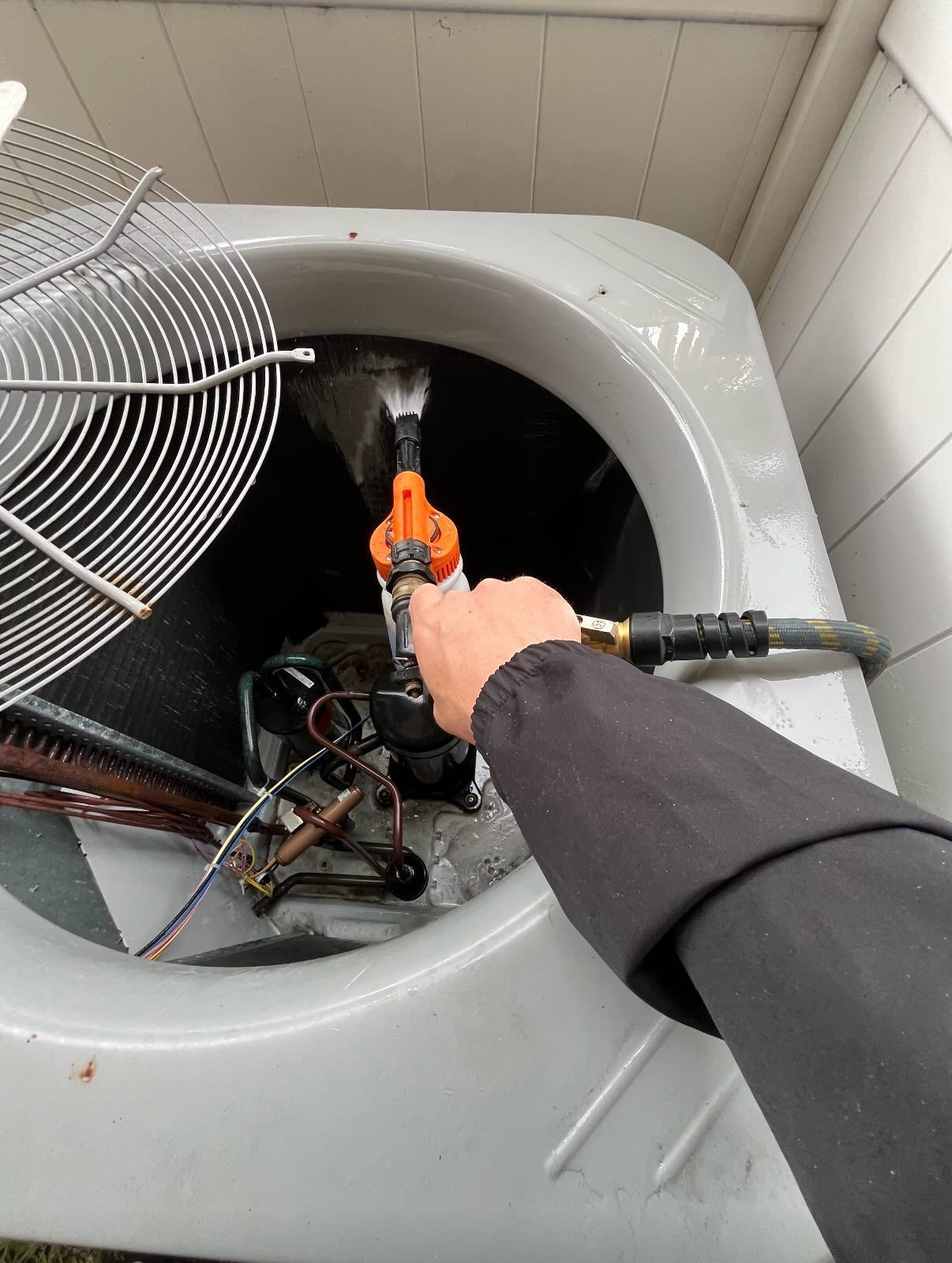 A hand uses a pressurized cleaning wand to spray water inside an outdoor HVAC condenser unit.