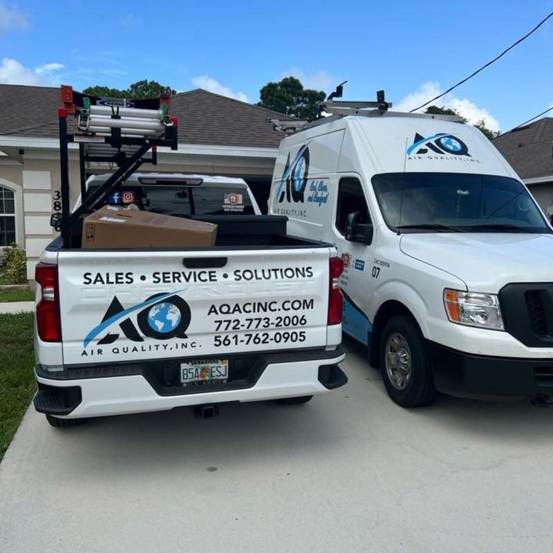 A white service pickup truck and a van parked in a driveway, both branded with the AQ Air Quality, Inc. logo and phone numbers.