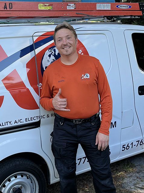 A technician wearing an orange long-sleeve shirt stands smiling in front of a white service van, making a shaka sign.