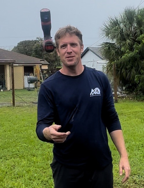 A person stands in a grassy yard, looking at the camera as a power drill floats in the air near their head.
