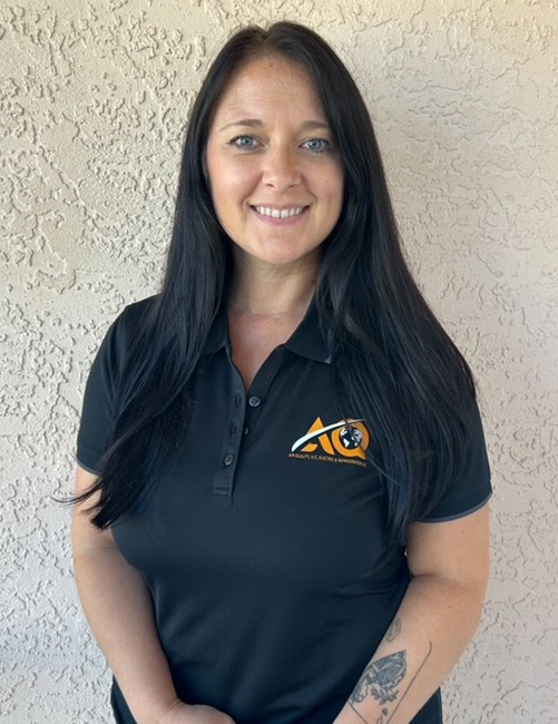 A smiling person with long dark hair wears a black collared shirt with an orange and white logo against a textured wall.