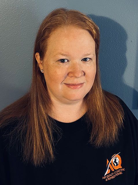 A person with long reddish-brown hair smiles gently in front of a blue wall, wearing a black shirt with a logo.