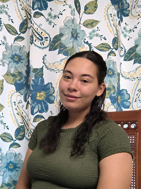 A person with dark hair sits in front of a blue and white floral curtain wearing a green textured top.
