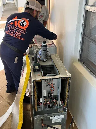 A technician in a navy uniform repairs an open HVAC unit in a hallway.