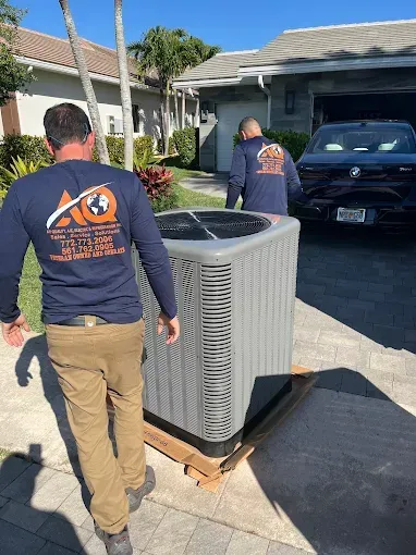 Two technicians in branded navy shirts push a new residential HVAC unit on a wooden pallet along a residential driveway.