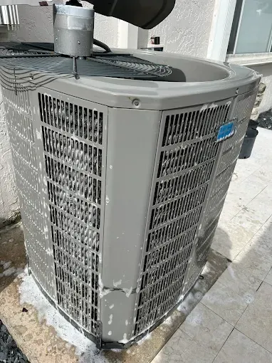 An outdoor residential air conditioning unit covered in white cleaning foam, sitting on a concrete patio.