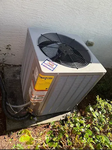 A gray outdoor air conditioning condenser unit sitting on a concrete pad next to a light-colored exterior wall.