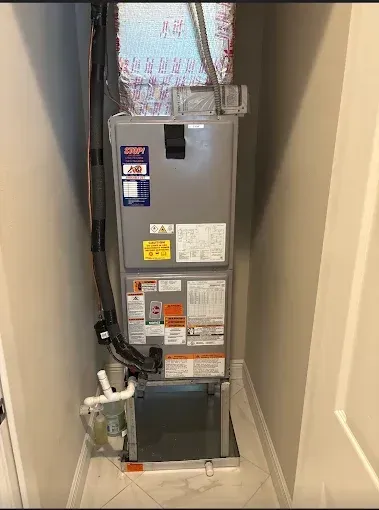 A grey HVAC air handler unit standing vertically inside a narrow indoor utility closet with white tile flooring.