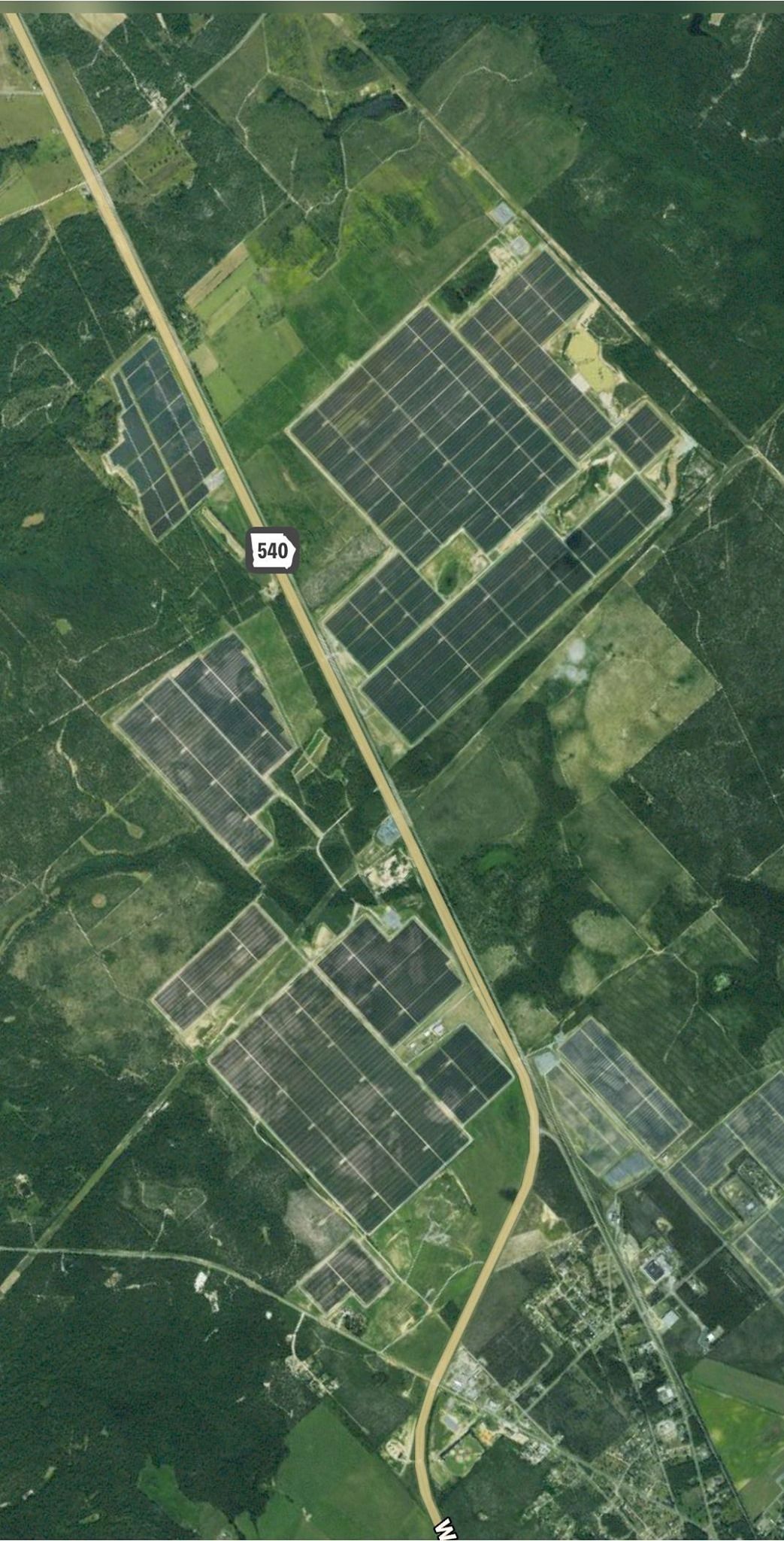 Aerial view of a solar panel farm along a highway in a rural area, featuring rectangular fields surrounded by trees.