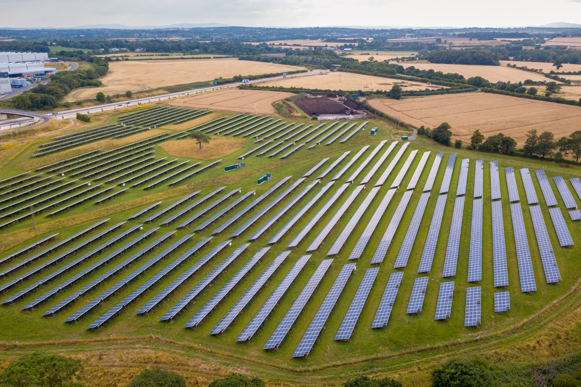 Aerial view of a solar farm on a grassy field; multiple rows of solar panels stand in neat rows, with farmland in the background.