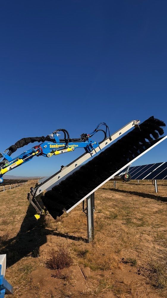 Three workers in orange coveralls clean solar panels on a rooftop with long-handled brushes.