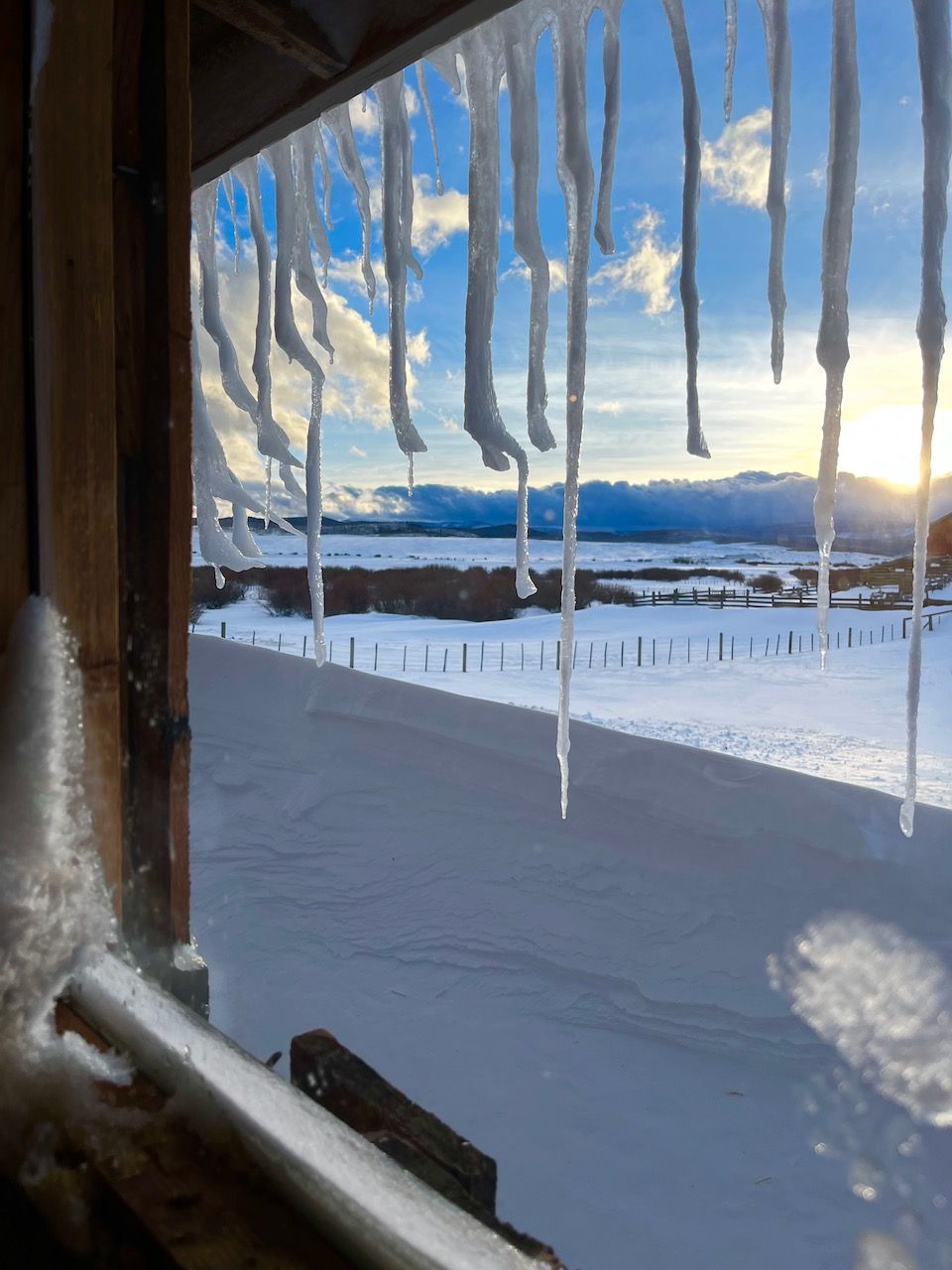 Icicles are hanging from a window overlooking a snowy field.