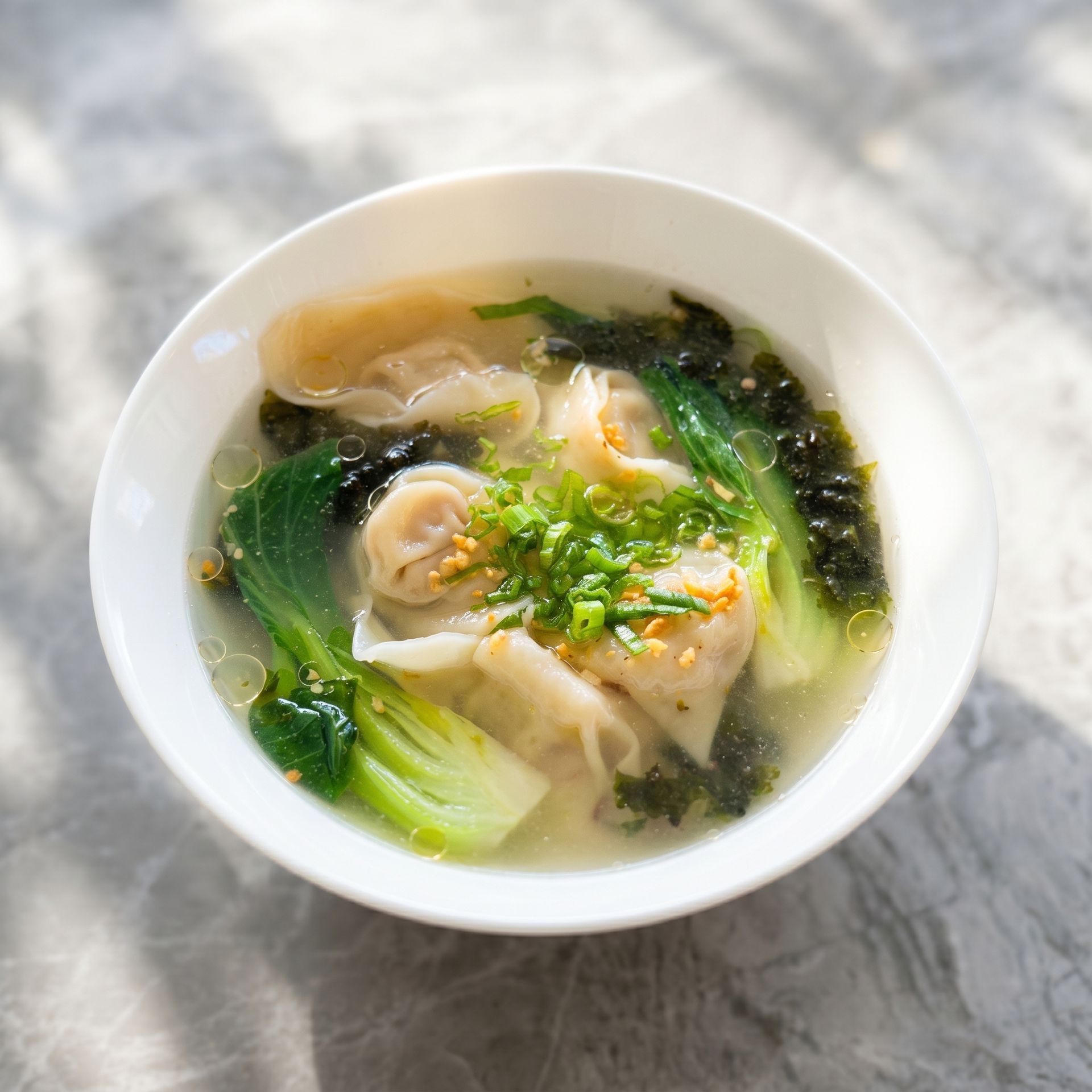 Bowl of clear noodle soup with shrimp dumplings, leafy greens, and chopped herbs on a marble table
