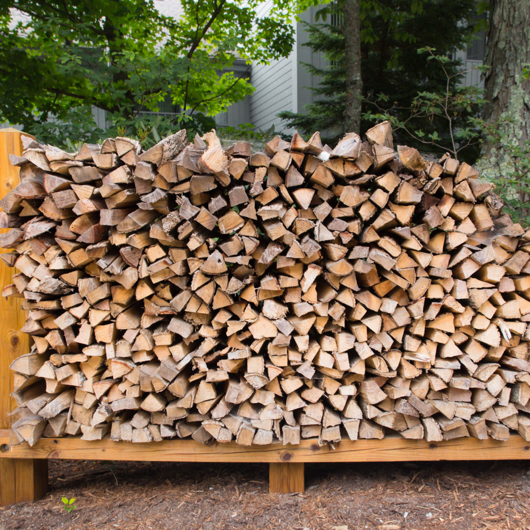 Pile of firewood stacked neatly on a wooden rack, set outdoors with trees and a building in the background.