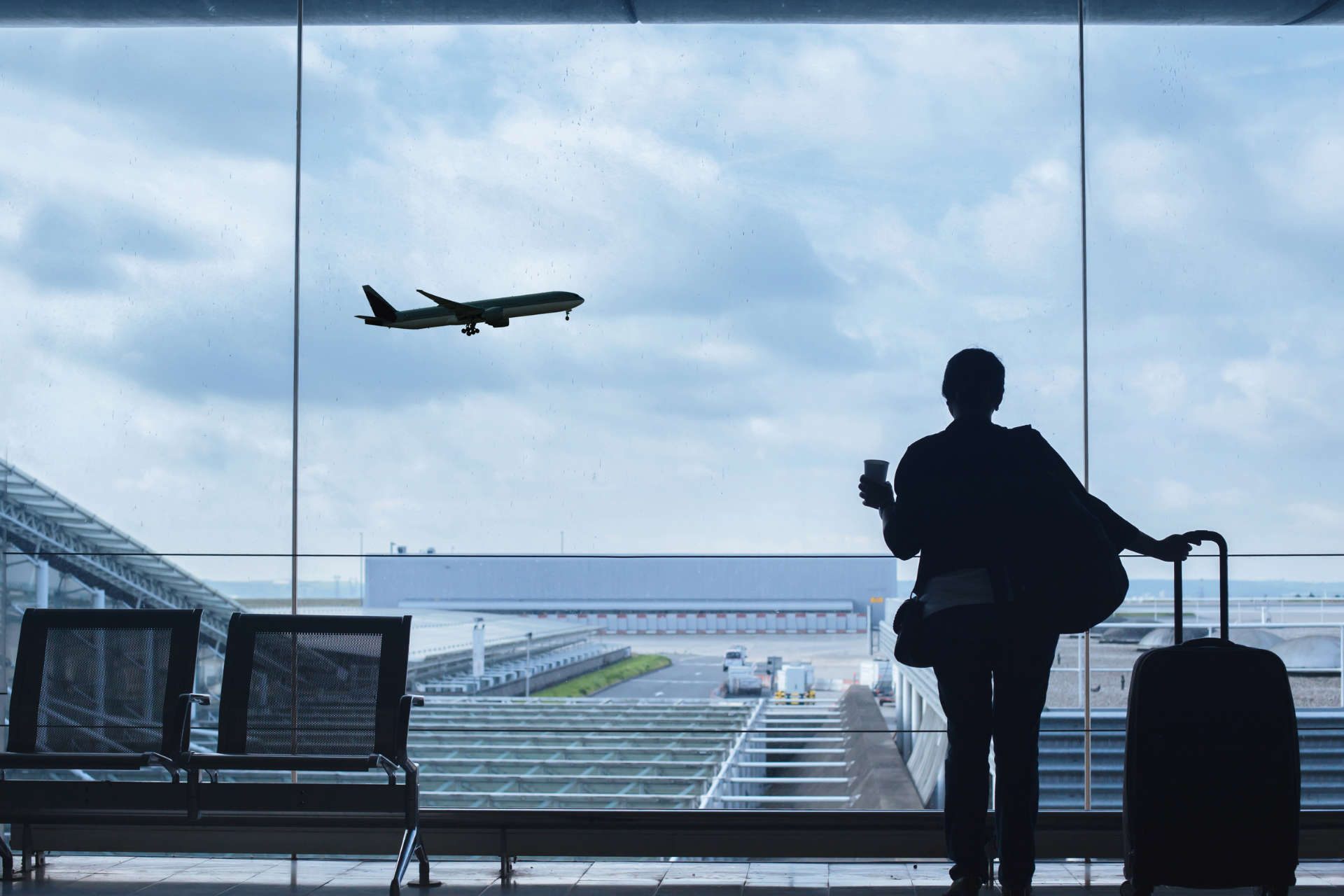 A man with a suitcase is looking out a window at an airplane taking off.