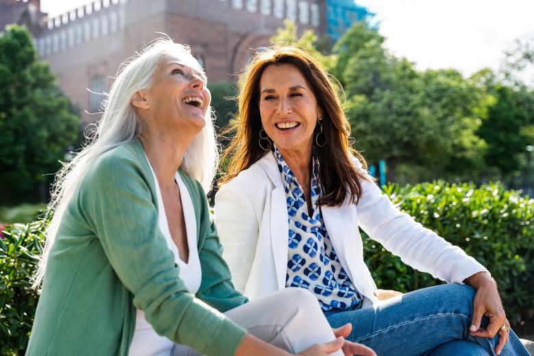 Two older women are sitting next to each other in a park and laughing.