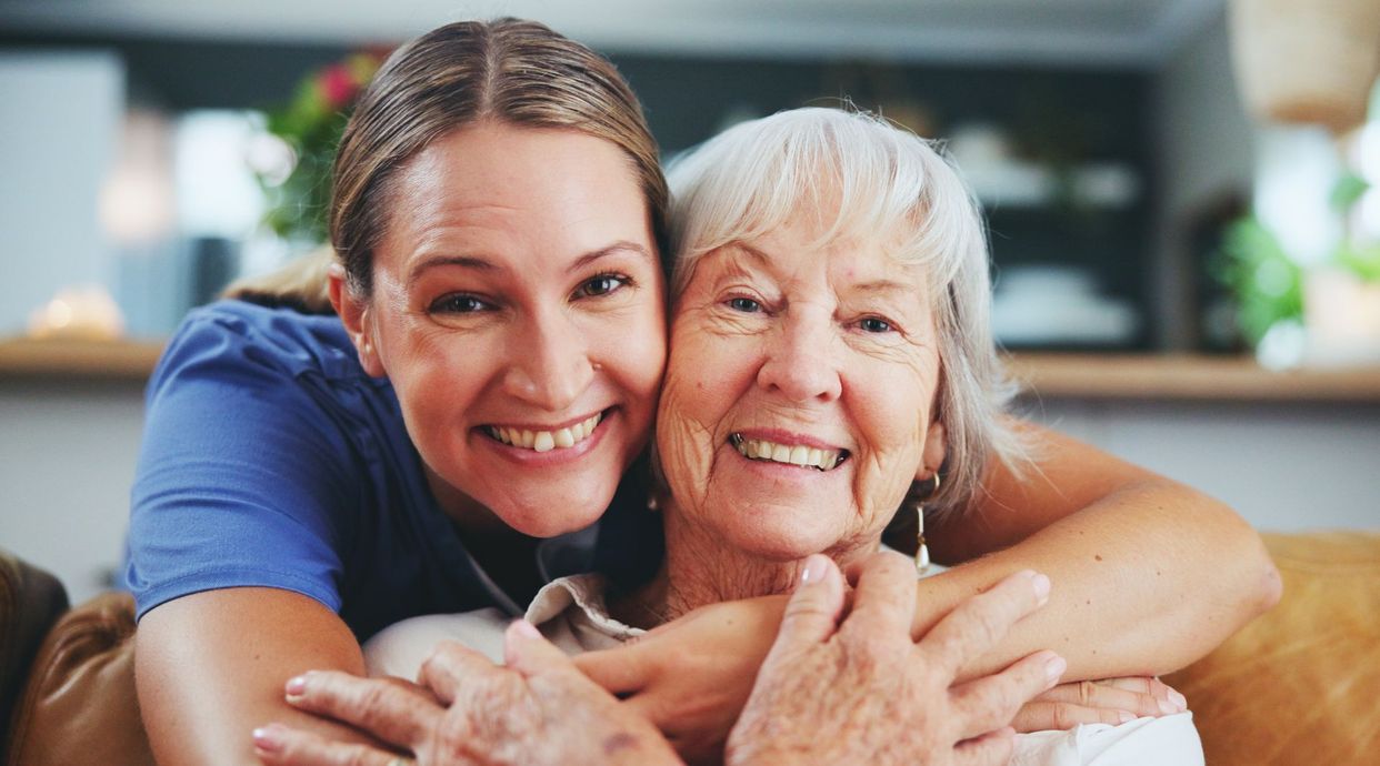 Woman hugging and smiling with an elderly woman; both are smiling and indoors.