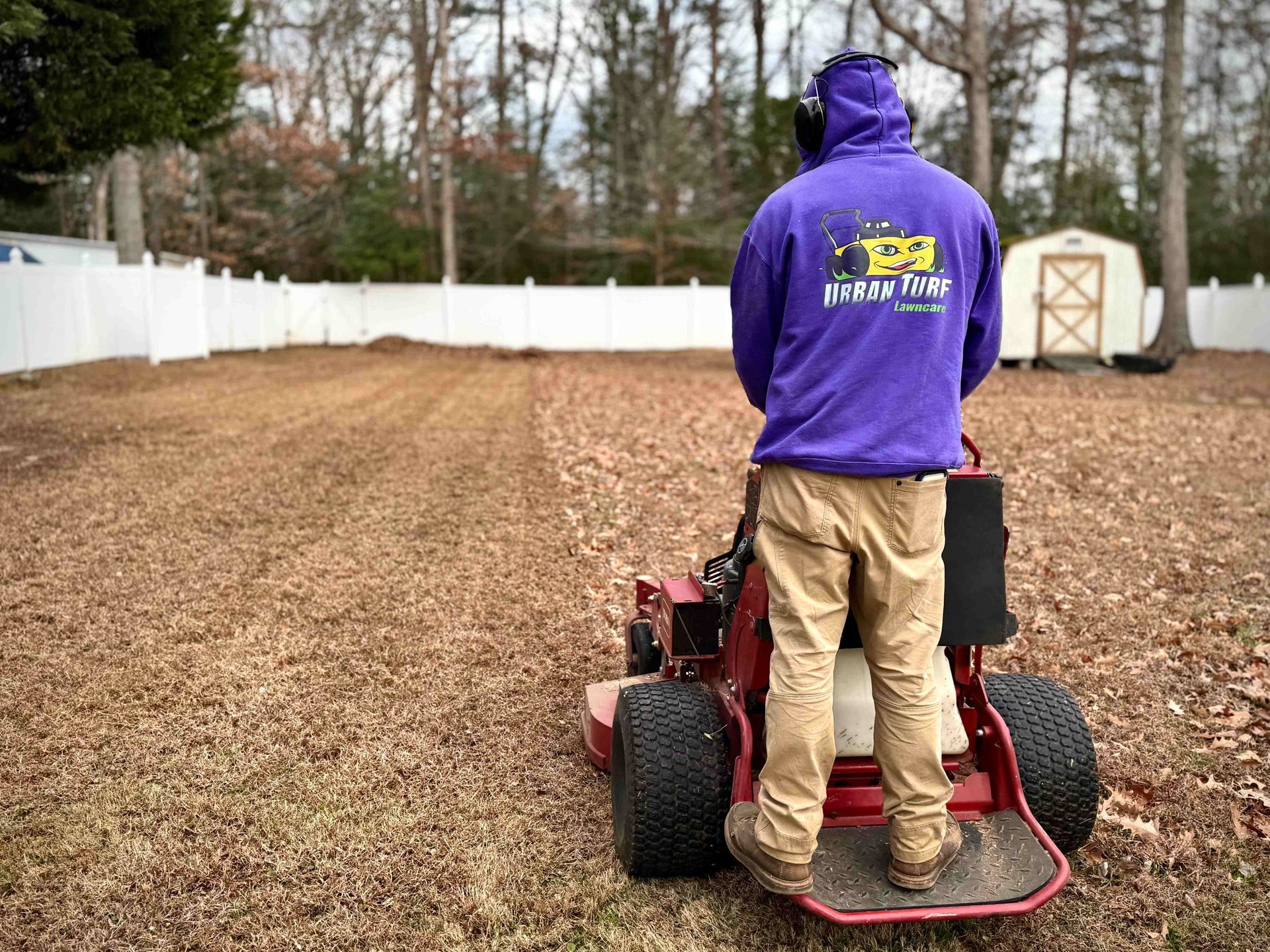 Person mowing a lawn with a riding mower, wearing a purple hoodie, khaki pants, and ear protection.