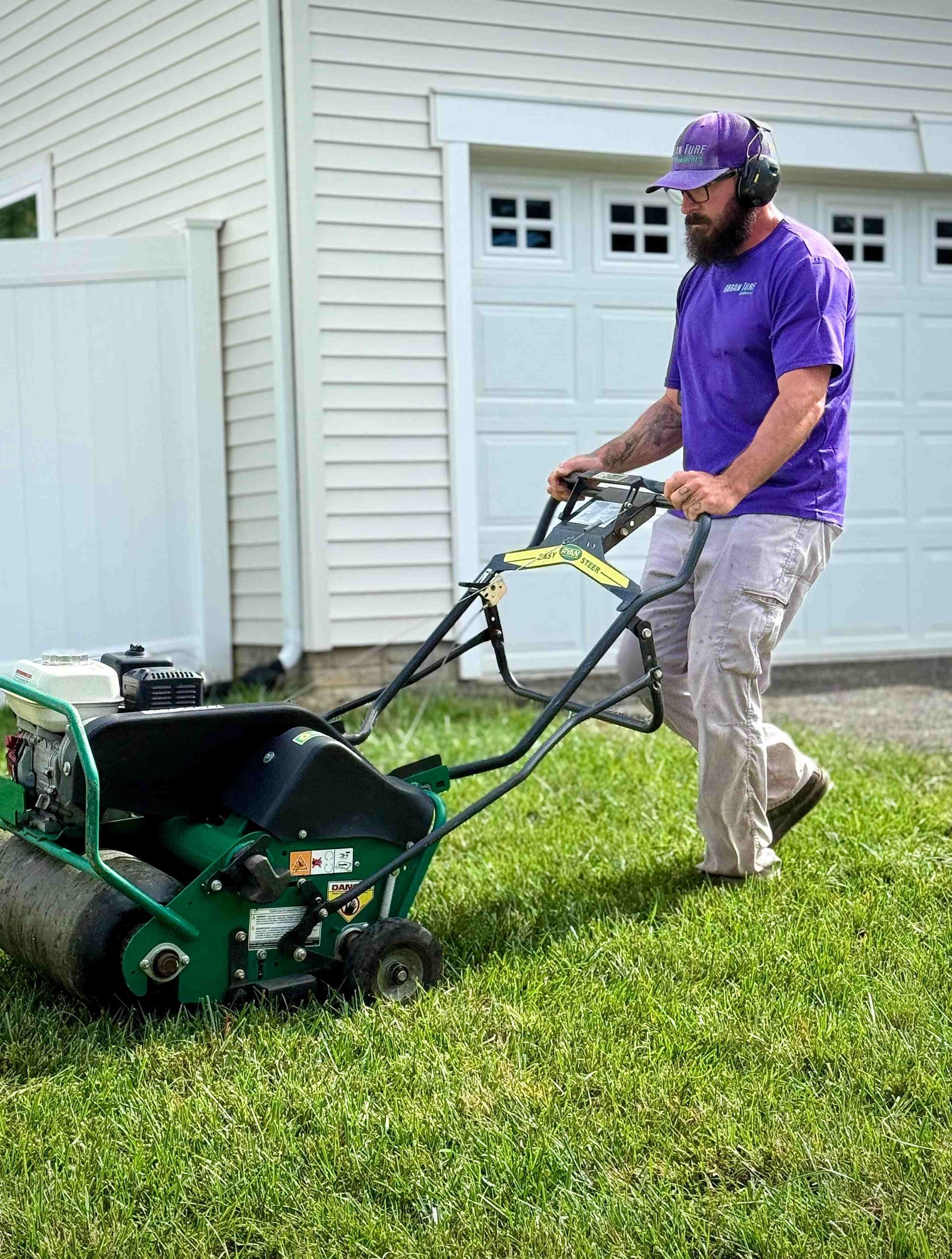 Man aerating lawn with machine near a white garage.
