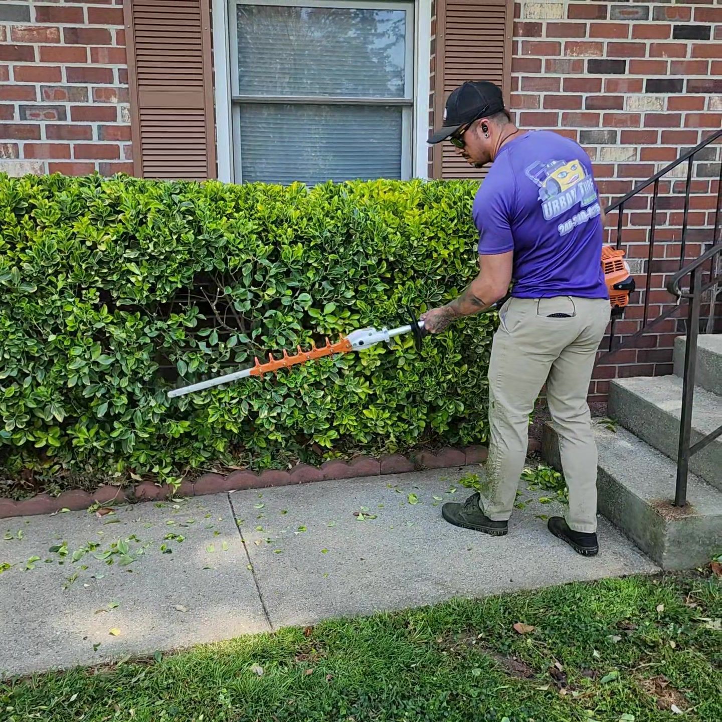 Man trimming hedge with electric shears next to a brick house.