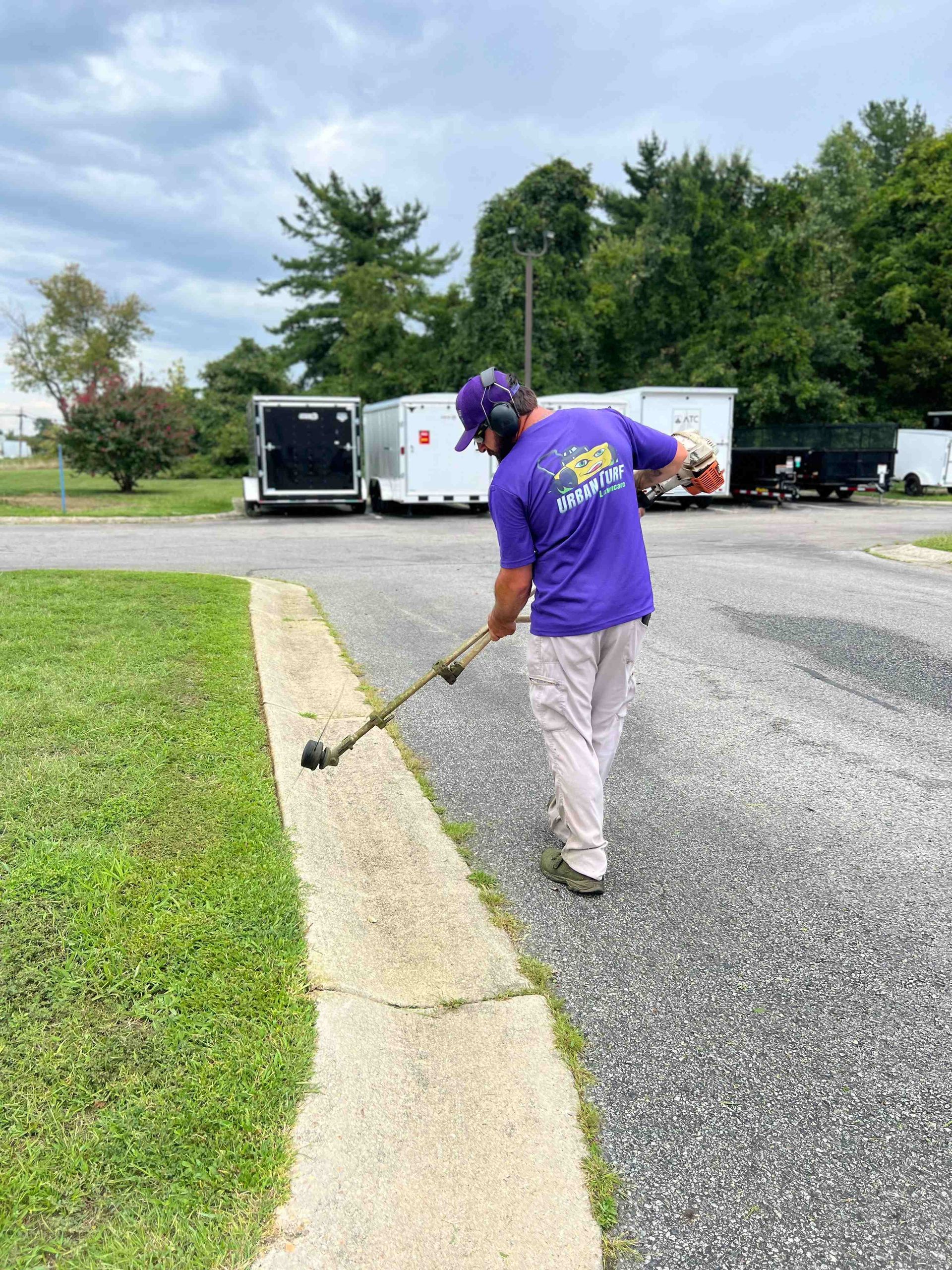 A person in purple shirt trims grass along a concrete edge with a weed whacker.