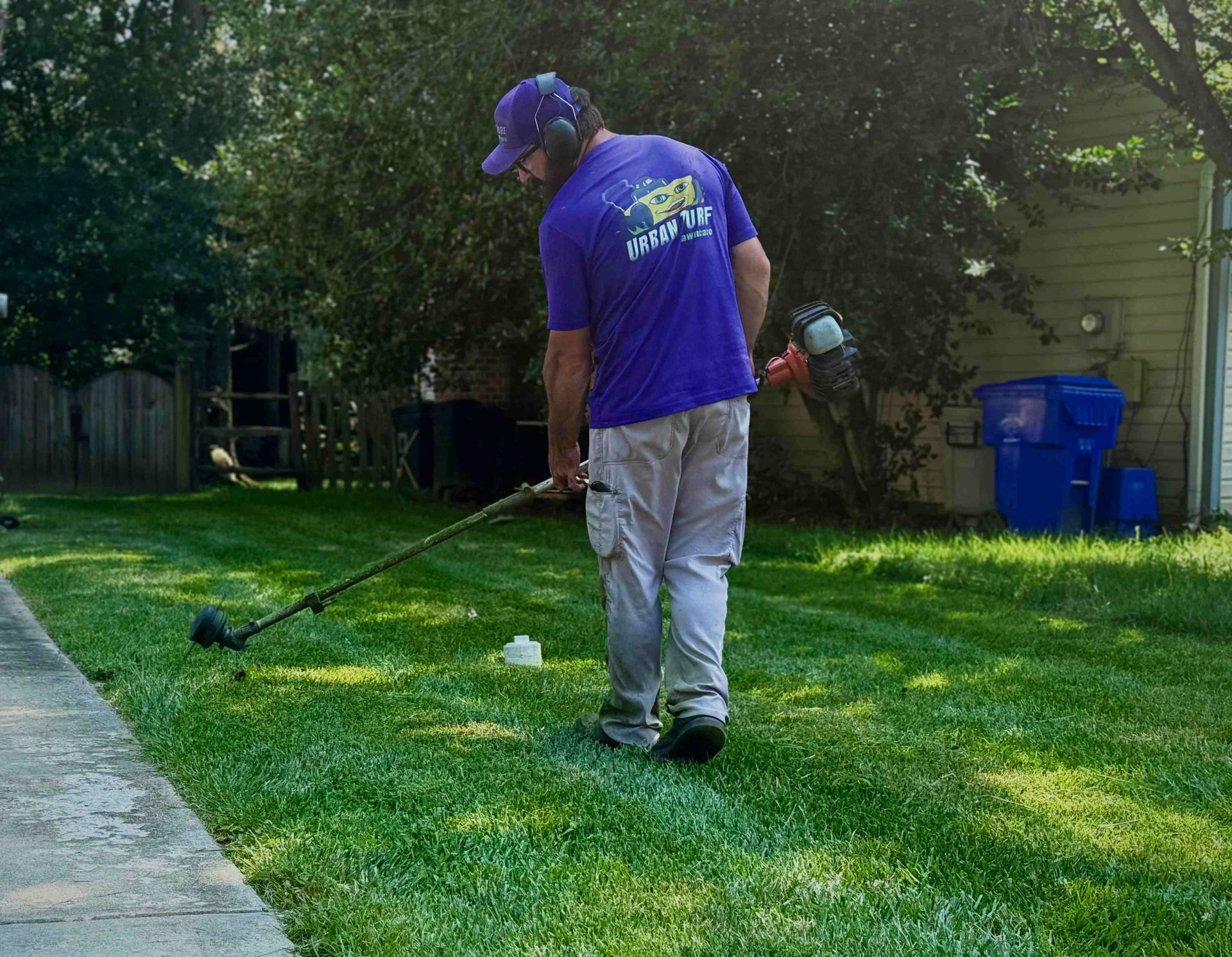 A person in a blue shirt uses a weed trimmer on a lawn next to a sidewalk.