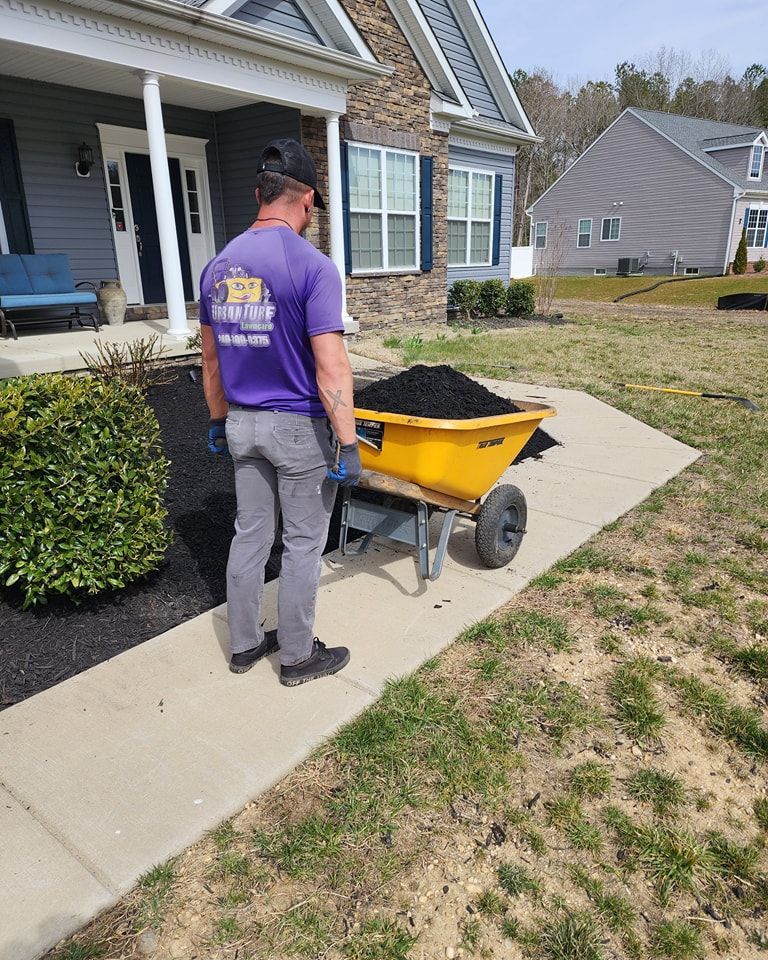 Man pushing a yellow wheelbarrow full of mulch on a sidewalk near a house with landscaping.