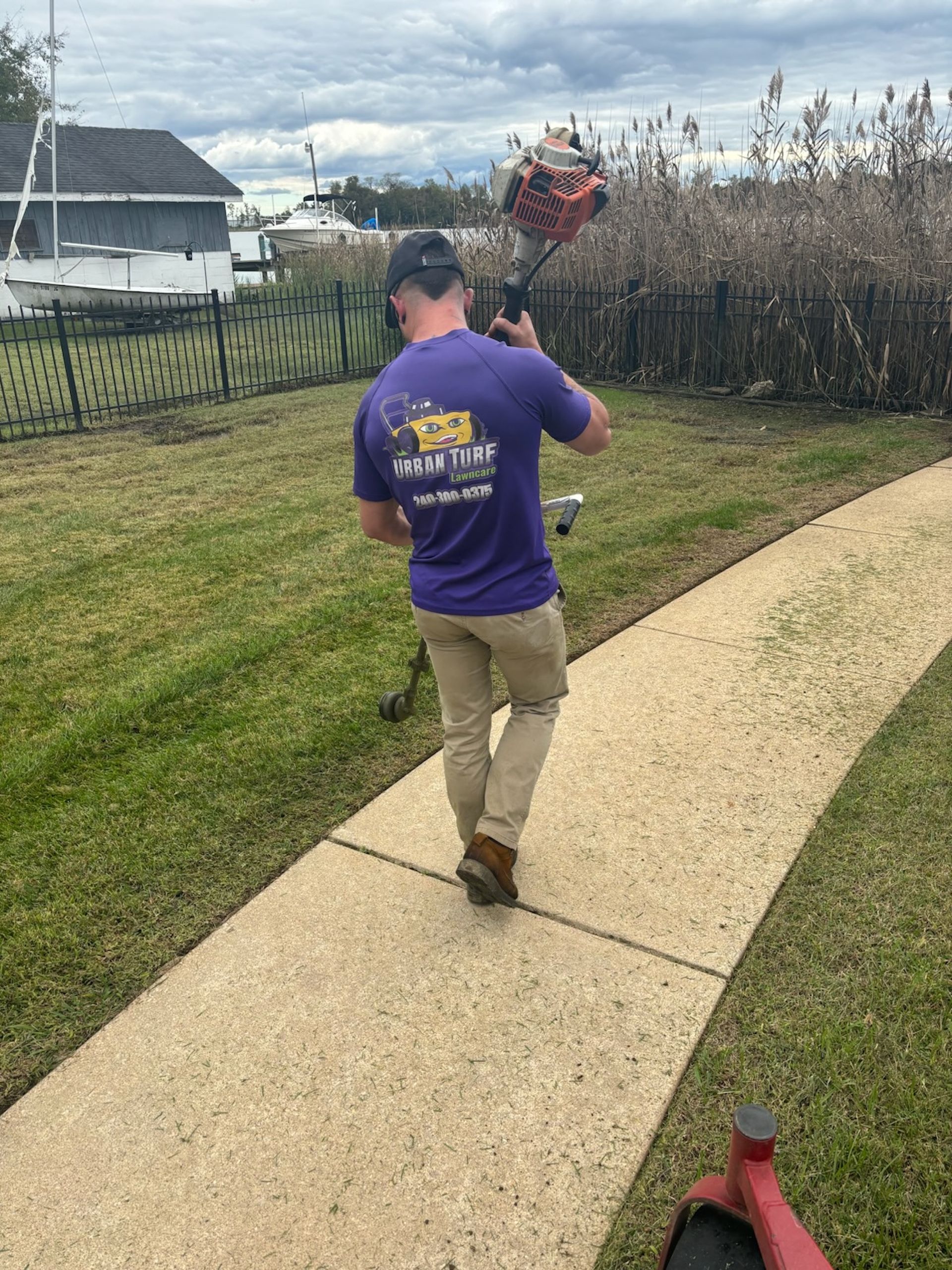 A man in a purple shirt is walking down a sidewalk holding a lawn mower.
