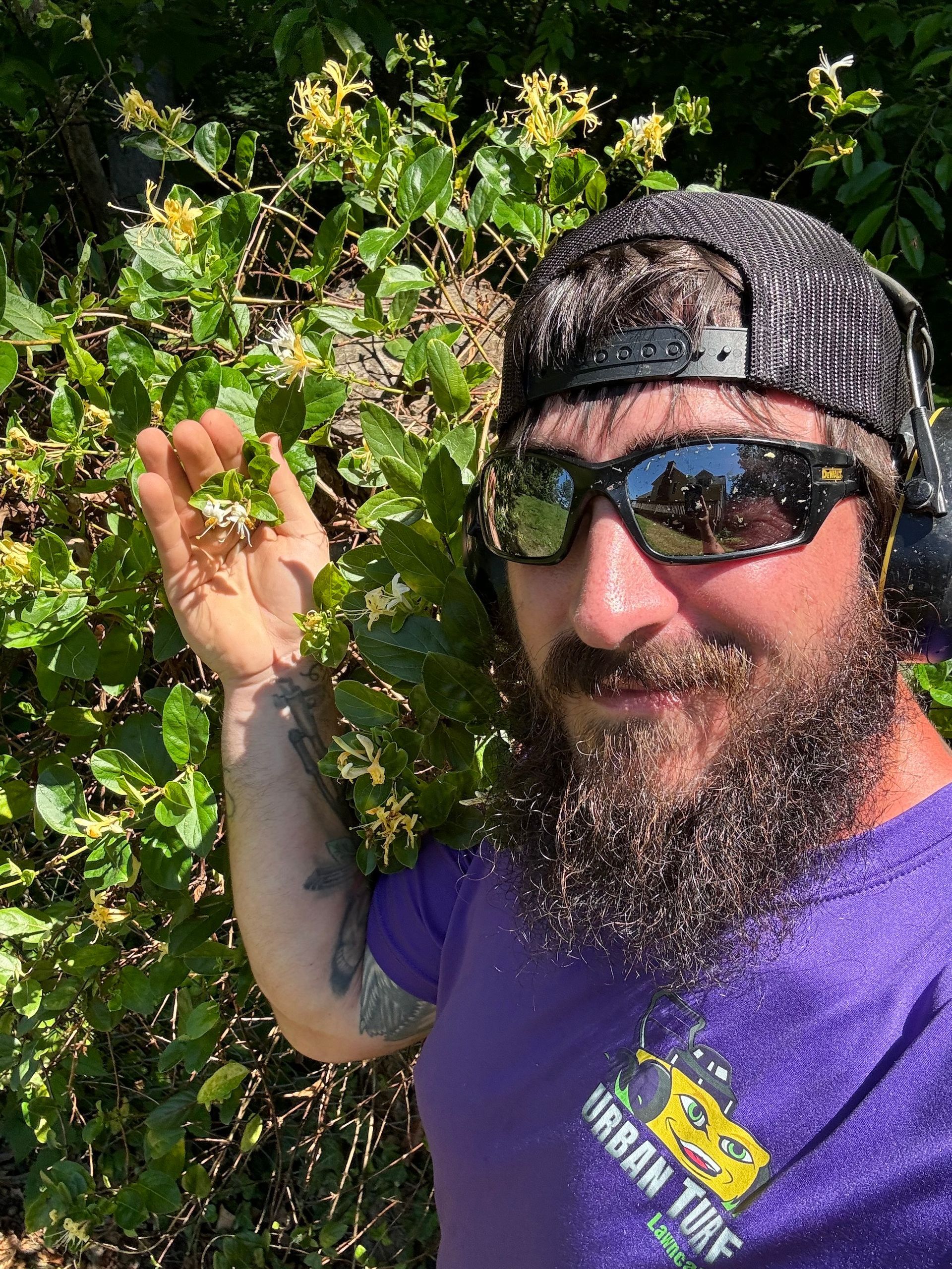 A man with a beard wearing sunglasses and headphones is standing in front of a bush.