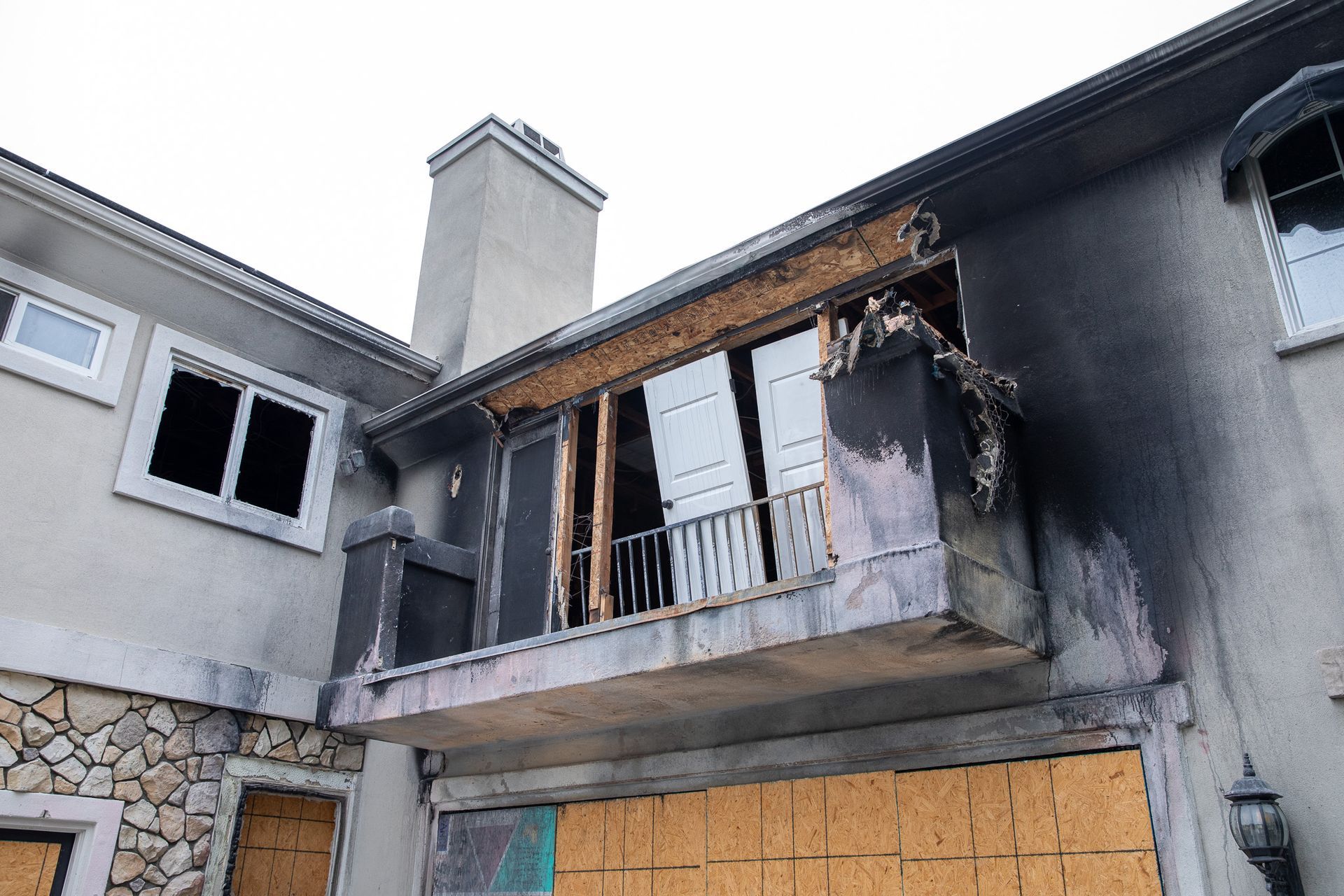 Exterior view of a house damaged by fire. Burned balcony, boarded windows, and soot are visible.