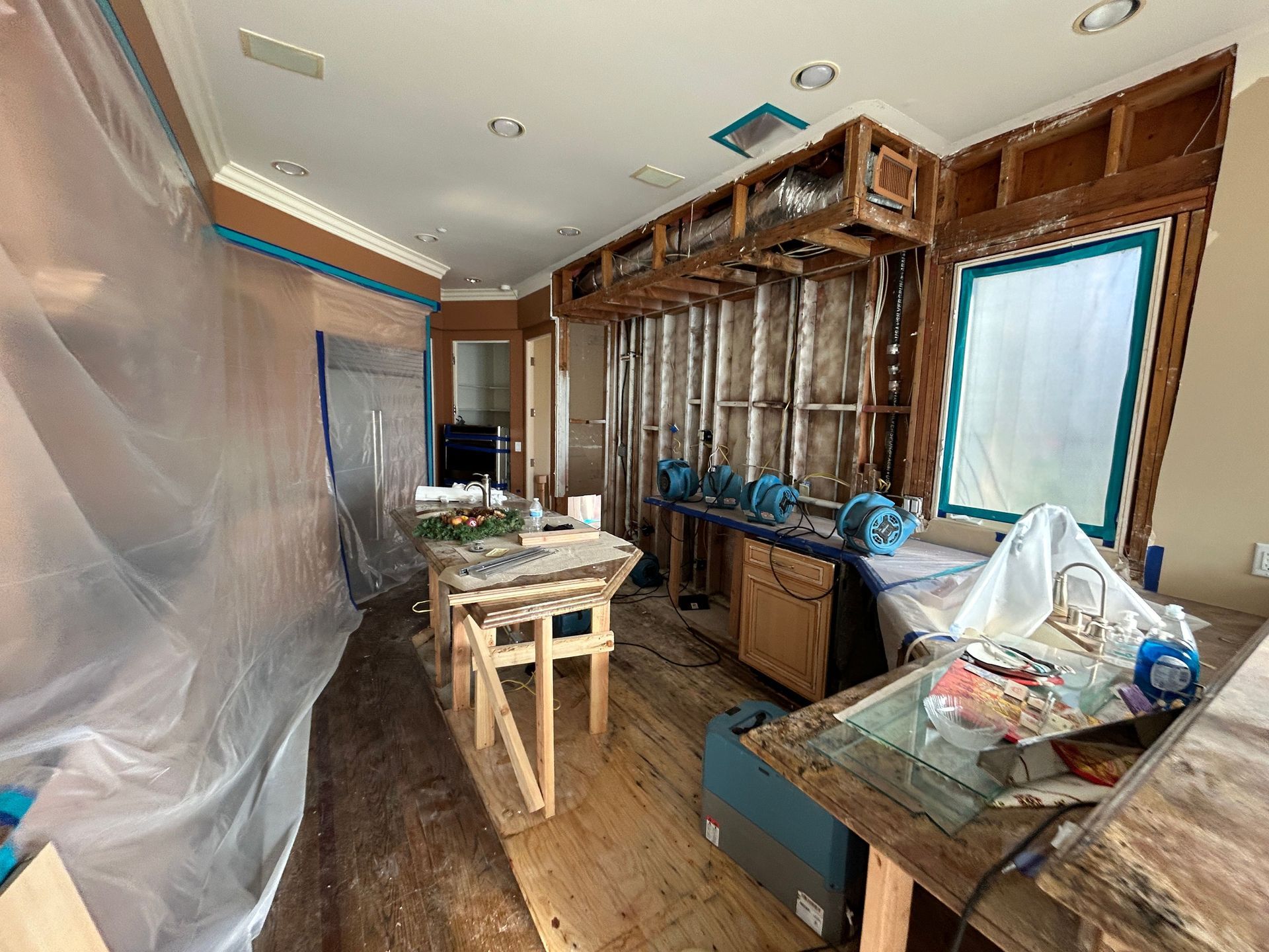 Kitchen renovation: walls open, exposed wood framing, plastic sheeting, tools, and fans.