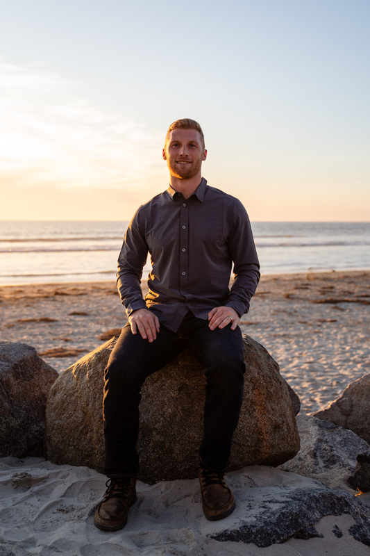 Man sitting on a rock at a beach, gazing at the sunset. He wears a button-up shirt and dark pants.